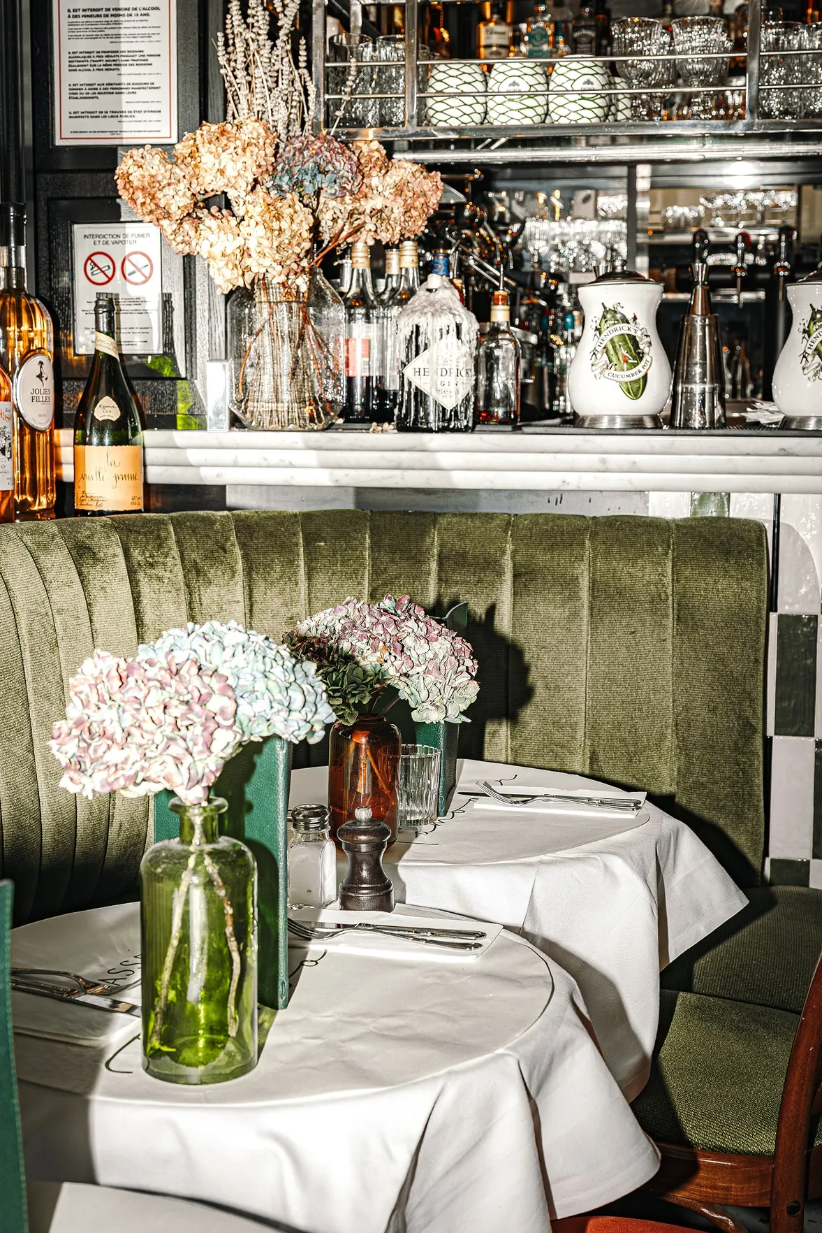 Cozy restaurant booth with green velvet seating, white tablecloths, vases of pastel hydrangeas, and a bar stocked with bottles and glassware in the background.