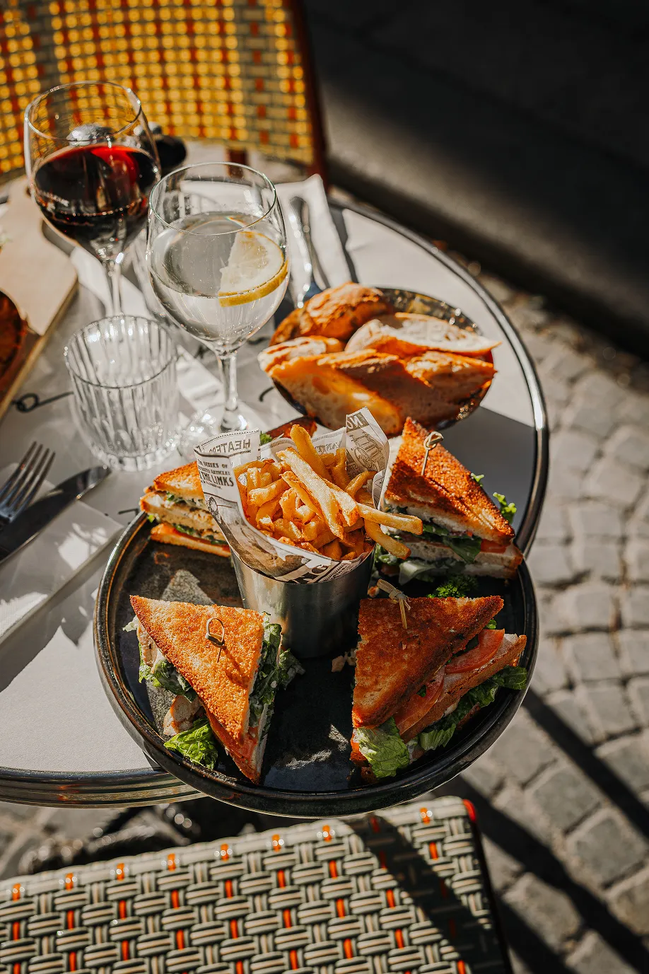 Plate with four triangular club sandwiches and a cup of French fries, accompanied by a bowl of bread slices and glasses of red wine and water with lemon on a round table outdoors.