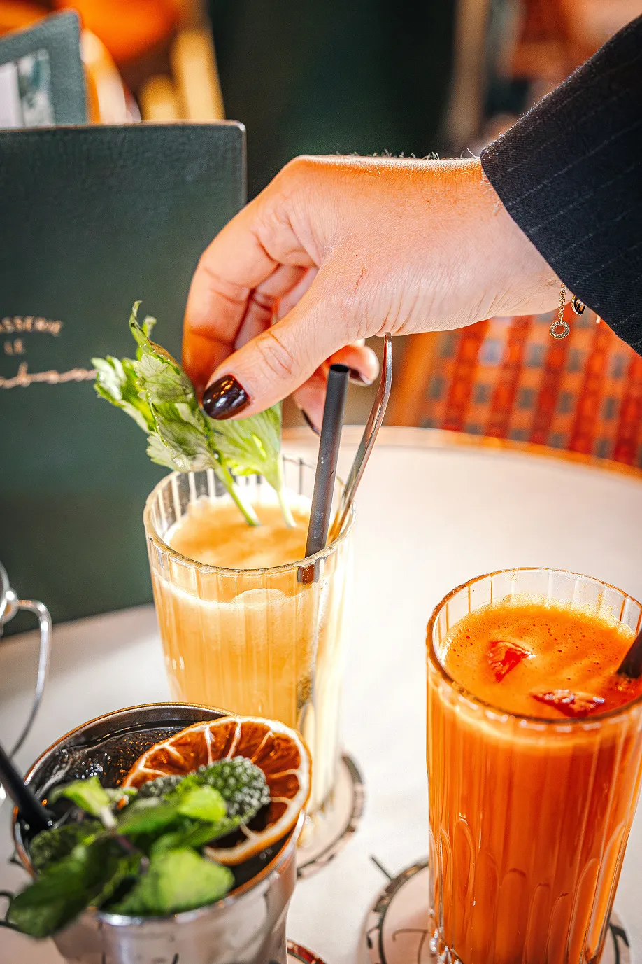 Hand with dark nail polish adjusting a straw in a peach-colored cocktail garnished with fresh mint leaves, next to two other colorful drinks on a table.